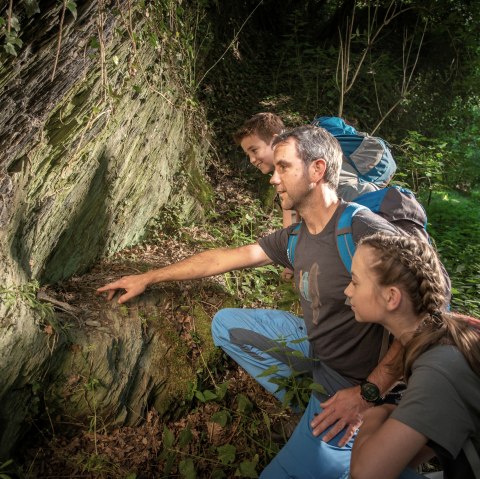 Drei Wanderer betrachten interessiert eine Felswand im Wald. Sie tragen Rucksäcke und sind von üppigem Grün umgeben., © kappest_remet Drei Wanderer betrachten interessiert eine Felswand im Wald. Sie tragen Rucksäcke und sind von üppigem Grün umgeben., © kappest_remet