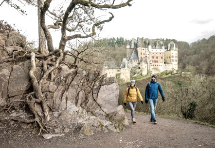 Zwei Wanderer in bunten Jacken gehen auf einem Pfad. Im Hintergrund ist die Burg Eltz zu sehen, umgeben von Wald., © Eifel Tourismus GmbH, Dominik Ketz Zwei Wanderer in bunten Jacken gehen auf einem Pfad. Im Hintergrund ist die Burg Eltz zu sehen, umgeben von Wald., © Eifel Tourismus GmbH, Dominik Ketz