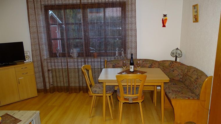 Ein gemütlicher Wohnbereich mit einem Esstisch und einer Sitzbank. Vor dem Fenster hängen Vorhänge, und es gibt eine Flasche Wein auf dem Tisch.