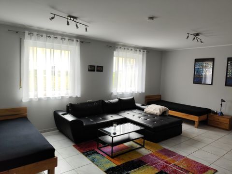Ein heller Wohnraum mit einem schwarzen Sofa und einladenden Fenstern. Der Raum ist mit einem bunten Teppich und minimalistischen Möbeln ausgestattet.