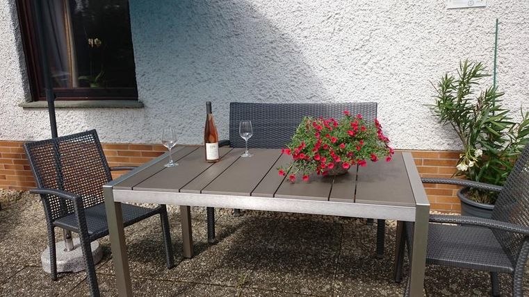 Ein schöner Tisch mit Stühlen auf einer Terrasse. Auf dem Tisch stehen eine Flasche Wein und zwei Gläser, umgeben von blühenden Pflanzen.