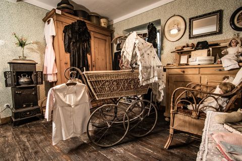 Ein nostalgisches Zimmer mit einem antiken Kinderwagen und historischem Mobiliar. An der Wand hängen alte Kleider, und ein Holzschrank steht im Hintergrund.