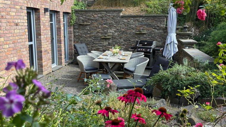Un beau jardin avec des fleurs colorées et un coin salon confortable. À l'arrière-plan, une maison moderne avec de grandes fenêtres est visible.