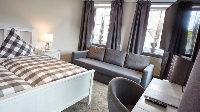 Ein modernes Schlafzimmer mit einem Bett, einem grauen Sofa und einem Arbeitsplatz. Große Fenster mit Vorhängen sorgen für viel Licht.