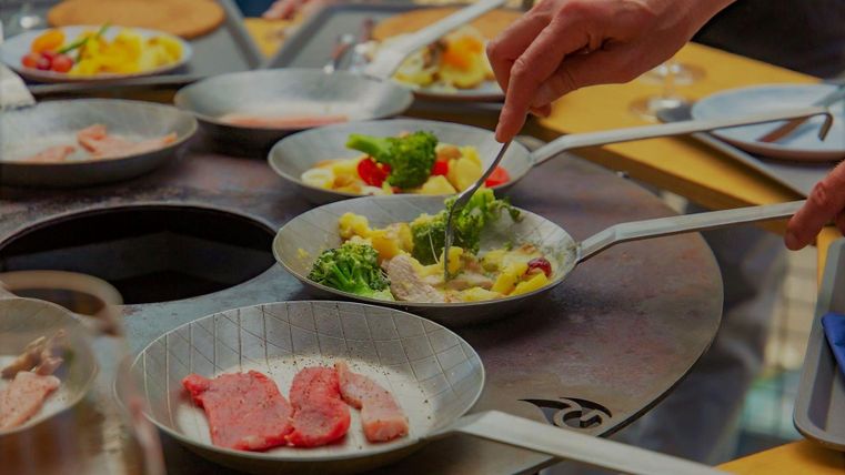Zwei Hände bereiten frische Zutaten in kleinen Pfannen vor. Es gibt Fleisch, Gemüse und eine bunte Auswahl an Beilagen.