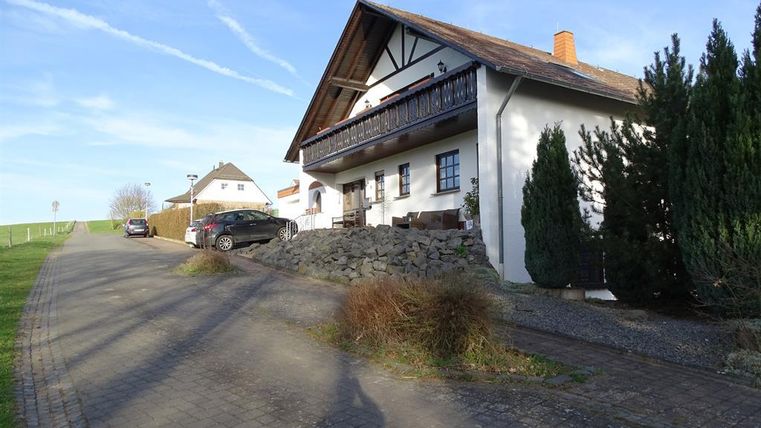 Ein modernes Haus an einer ruhigen Straße mit einem gepflasterten Weg. Es gibt Bäume und andere Häuser in der Umgebung.