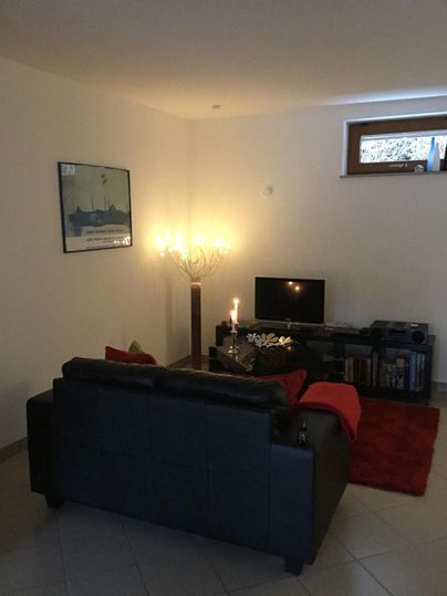 Ein gemütliches Wohnzimmer mit einem schwarzen Sofa, einem Fernseher und einem stilvollen Lampenständer. Es gibt eine warme Atmosphäre durch Kerzenlicht und eine rote Decke.