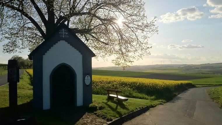 Eine kleine Kapelle steht auf einem Hügel, umgeben von grünen Feldern und einem Baum. Die Sonne scheint im Hintergrund und zaubert eine friedliche Atmosphäre.