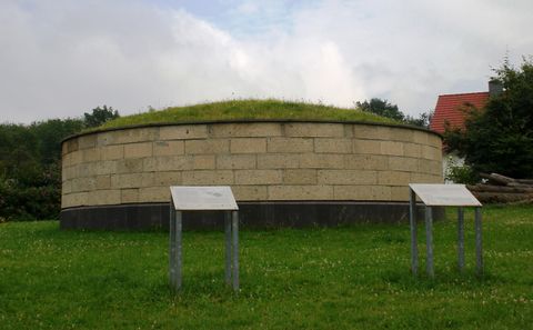 Ein runder, gemauerter Hügel mit einer grünen Grasbedeckung. Es stehen zwei Informationsschilder davor.