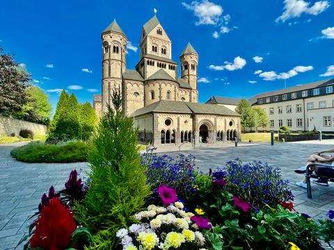 Eine beeindruckende Kirche mit Türmen und einem klaren Himmel. Im Vordergrund blühen bunte Blumen in einem gepflegten Garten.