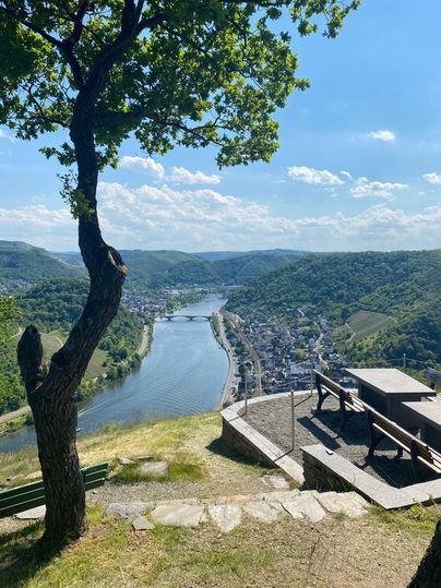 Eine malerische Aussicht auf einen Fluss, umgeben von grünen Hügeln und einem charmanten Dorf. Der Himmel ist blau mit einige Wolken.