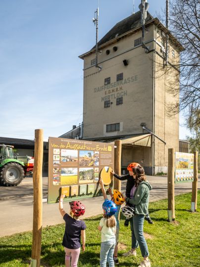Eine Mutter mit Kindern schaut die Infotafel zur Landwirtschaft am Maifeld-Radweg an