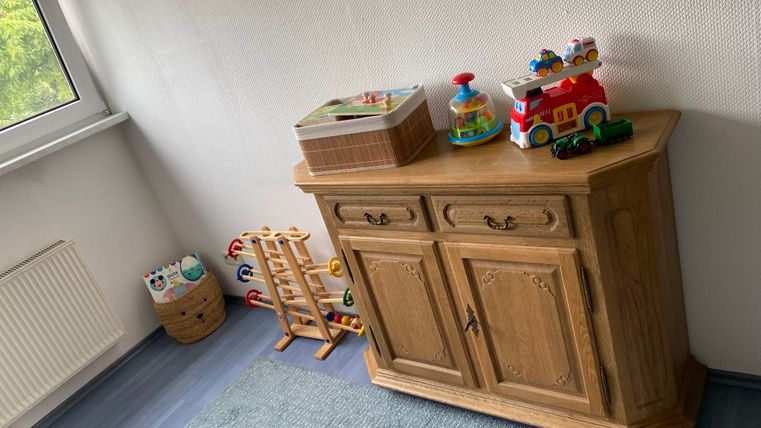 Ein heller Raum mit einem Holzschrank. Auf dem Schrank stehen Spielzeuge und ein paar Spielsachen sind auf dem Boden zu sehen.