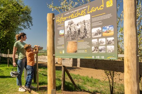 Eine Mutter steht mit ihrem Kind vor einem Informationsschild über historisches Land. Die Umgebung ist grün und sonnig.