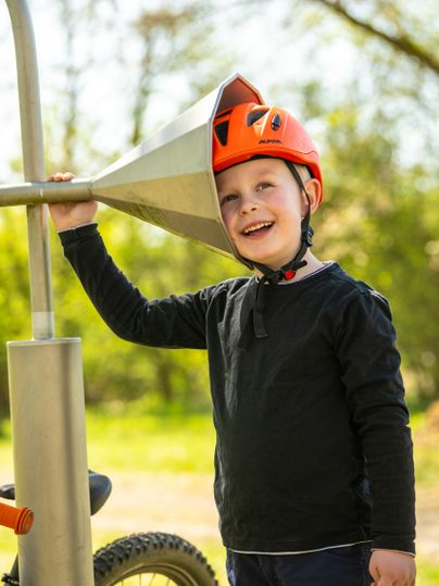 Ein Junge mit Fahrradhelm probiert das Dosentelefon auf dem Sinnespfad am Maifeld-Radweg