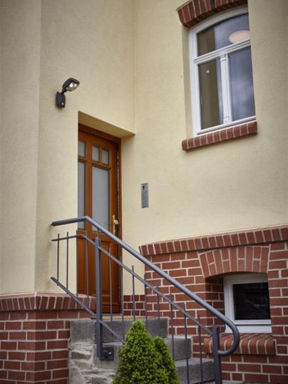 Une entrée avec une porte brune et une grande fenêtre. Un escalier en métal mène à la porte d'entrée, entourée d'un mur clair et de briques rouges.