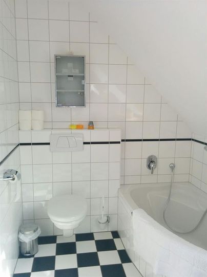 Ein einfaches, modernes Badezimmer mit weißen Fliesen und einem schwarzen und weißen Schachbrettmuster auf dem Boden. Es enthält eine Badewanne, eine Toilette und einen Spiegel.