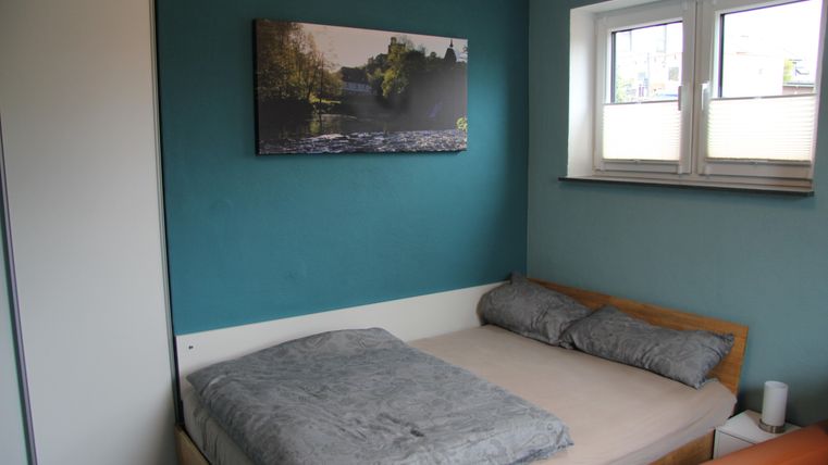 Une chambre confortable avec un lit double et du linge de lit gris.
Sur le mur, il y a une image d'un paysage, et il y a une fenêtre laissant entrer la lumière naturelle.