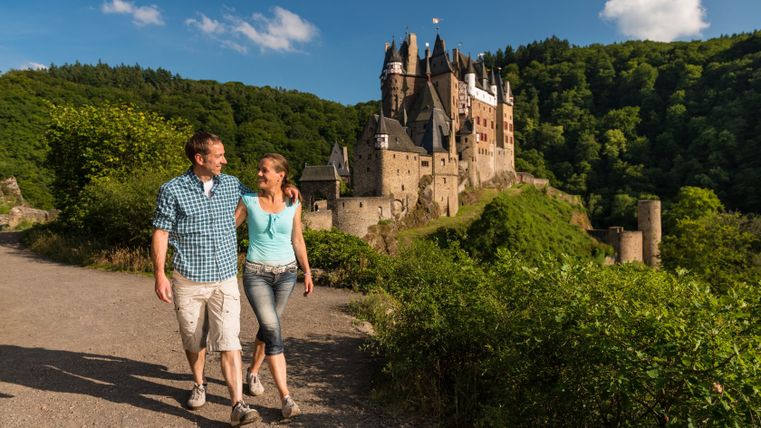 Ein Paar spaziert auf einem Weg mit Burg Eltz im Hintergrund.