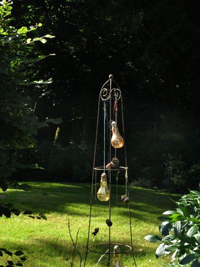 Ein dekorativer Metallständer mit gläsernen Kugeln steht in einem grünen Garten. Umgeben von Pflanzen und Bäumen, dringt sanftes Licht durch das Laub.