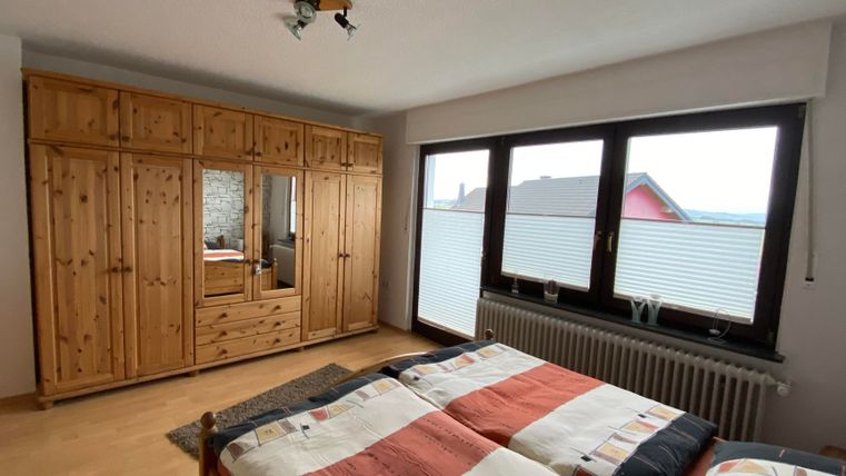 Ein gemütliches Schlafzimmer mit einem Doppelbett und einem großen Kleiderschrank aus Holz. Die Fenster bieten einen Blick nach draußen und lassen viel Licht herein.