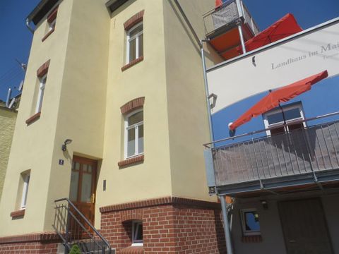 Un bâtiment jaune avec une maçonnerie en brique rouge et un escalier à l'entrée. Sur le balcon, on voit des parasols rouges.