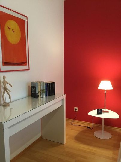 Ein gemütliches Zimmer mit einem weißen Schreibtisch, einem Kunstwerk an der Wand und roten Akzenten. Eine Lampe und einige Bücher verleihen dem Raum eine einladende Atmosphäre.