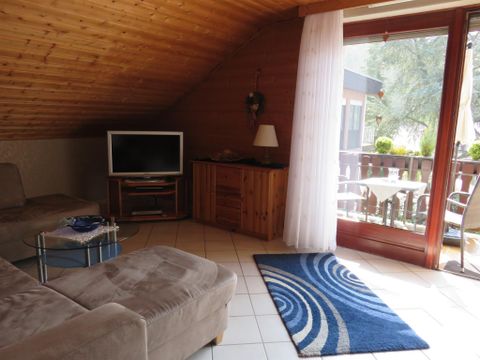 Ein gemütlicher Wohnraum mit einem großen Sofa und einem Fernseher. Die Fenster bieten einen Blick auf einen Balkon mit Gartenmöbeln.