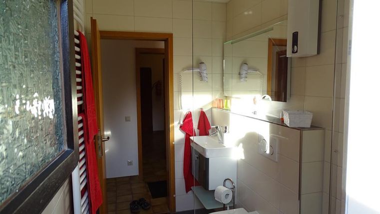 Ein helles Badezimmer mit modernen Armaturen und einem großen Spiegel. Auf der linken Seite hängen rote Handtücher.