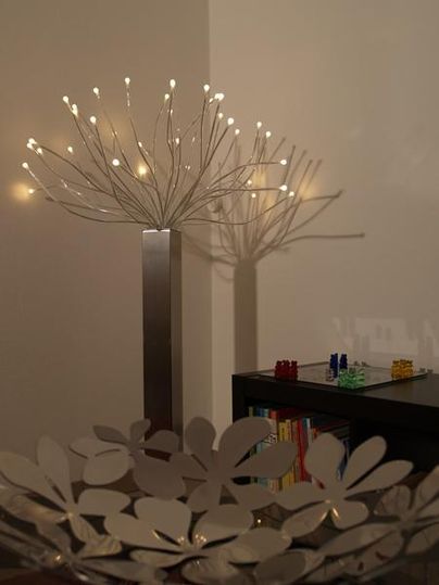 Eine moderne Lampe in Form eines baumähnlichen Designs steht an der Wand und wirft einen Schatten. Im Vordergrund ist eine dekorative Schale mit blütenförmigen Motiven sichtbar.