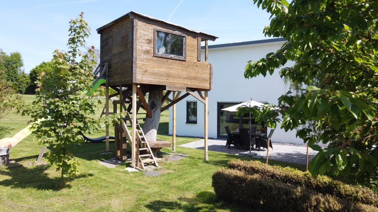 Une maison de jeu en bois sur pilotis dans un jardin. À côté, il y a une table avec des chaises et un parasol.