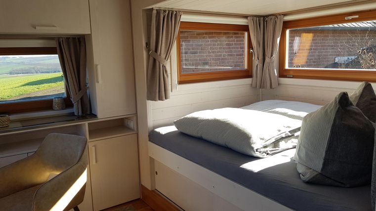 Ein helles Zimmer mit einem Bett, großen Fenstern und einem gemütlichen Sessel. Der Blick nach draußen zeigt eine grüne Landschaft.