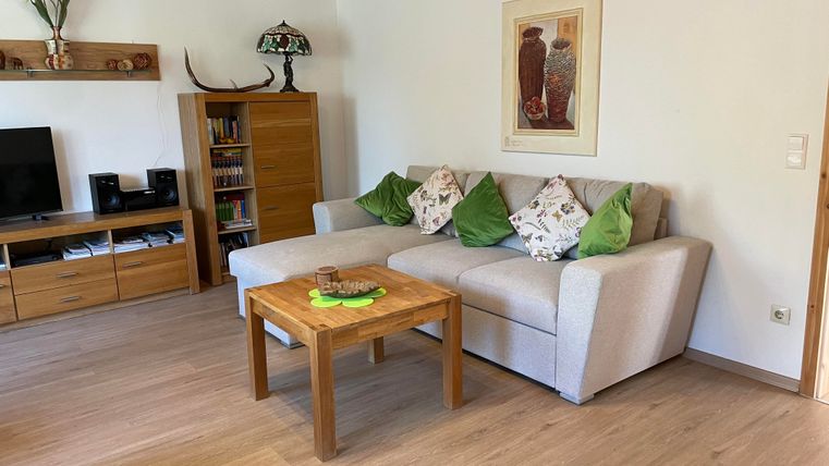 Ein gemütliches Wohnzimmer mit einem hellen Sofa, dekorierten Kissen und einem Holztisch. An der Wand hängen Bilder, und im Hintergrund steht ein Fernseher.