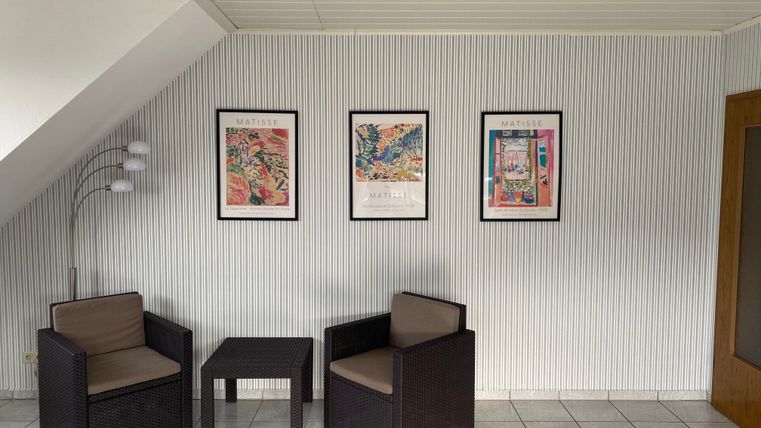 Ein heller Raum mit zwei Stühlen und einem kleinen Tisch. An der Wand hängen drei gerahmte Bilder.