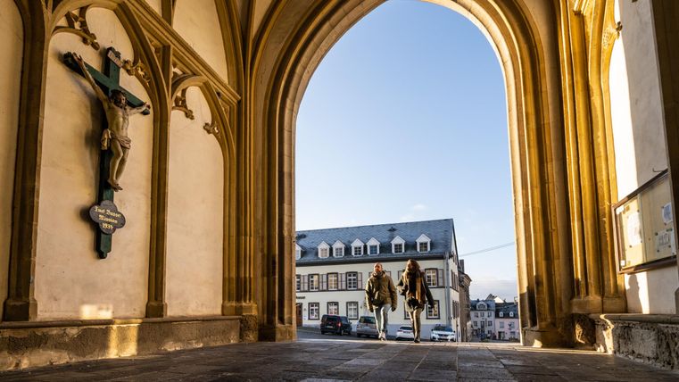 Ein Blick durch ein großes Tor mit gotischen Bögen auf eine Straße und ein historisches Gebäude. Zwei Personen gehen zusammen in die Ferne.