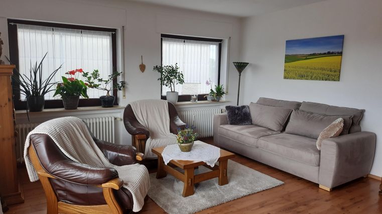 Ein gemütliches Wohnzimmer mit zwei bequemen Sesseln und einem Sofastuhl. Es gibt Pflanzen an den Fenstern und ein Bild an der Wand.