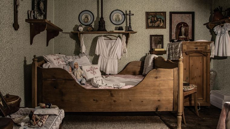 Ein gemütliches Schlafzimmer mit einem Holzbett und nostalgischen Dekorationen. An den Wänden hängen Bilder und religiöse Symbole.