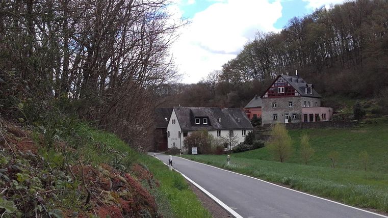 Een rustige landweg leidt langs traditionele huizen. De omgeving is groen en omgeven door bomen, met een heldere lucht.