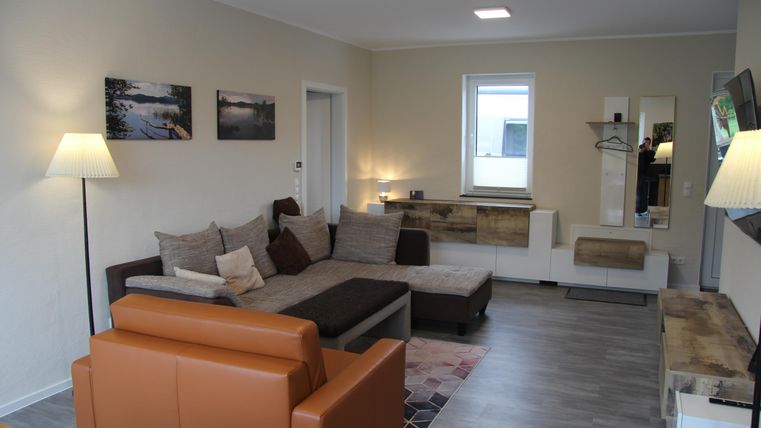 Un salon moderne avec un canapé confortable, un fauteuil brun et des murs clairs. Il y a des tableaux décoratifs sur le mur et un bon éclairage.