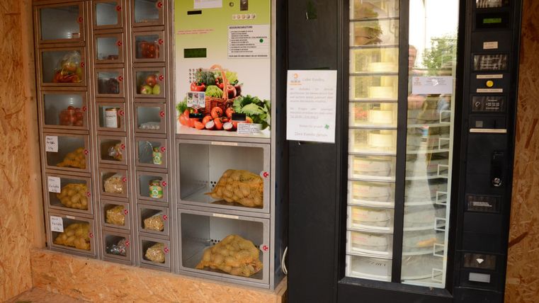 Ein Verkaufsautomat mit verschiedenen Snacks und ein weiterer Automat mit frischem Obst und Gemüse. Beide befinden sich in einem Holzrahmen.