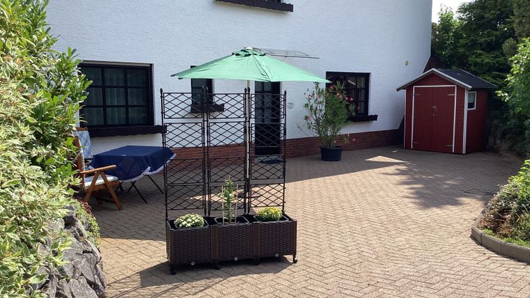 Ein schöner Innenhof mit einem Pflanzengitter und einem grünen Sonnenschirm. Im Hintergrund ist ein rotes Gartenhaus zu sehen.