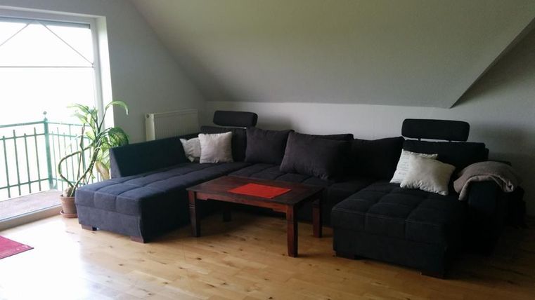 Ein gemütliches Wohnzimmer mit einer großzügigen Couch und einem Holztisch. Große Fenster lassen viel Tageslicht herein und bieten einen Blick nach draußen.