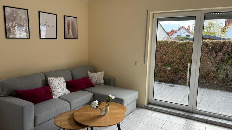 Ein gemütliches Wohnzimmer mit einem grauen Sofa und roten Kissen. Große Fenster bieten Blick auf einen begrünten Außenbereich.