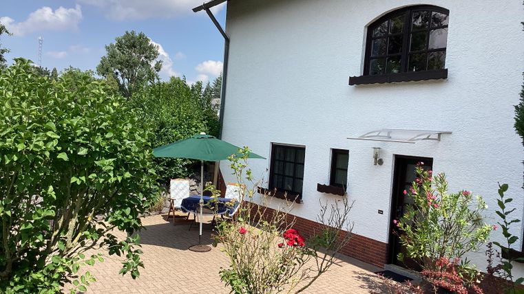 Ein hübsches Haus mit einem gepflegten Garten. Auf der Terrasse stehen Stühle und ein Sonnenschirm.