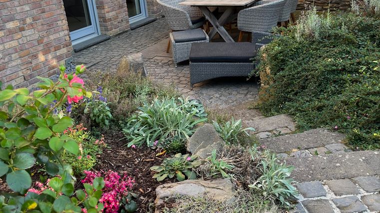 Un espace extérieur confortable avec une table et des chaises. Des fleurs colorées entourent la zone de sièges et un mur en pierre forme l'arrière-plan.