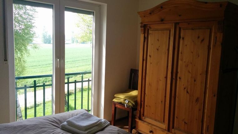 Ein helles Zimmer mit einem großen Fenster, das auf eine grüne Landschaft zeigt. Ein mittelgroßer Holzschrank und ein Stuhl sind im Raum sichtbar.