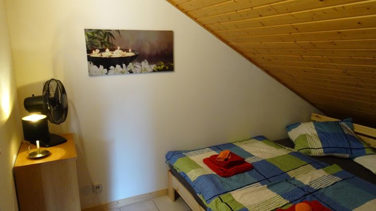 Ein gemütliches Schlafzimmer unter dem Dach mit einem Bett und bunten Decken. Auf einem Nachttisch steht eine Lampe und ein Ventilator.