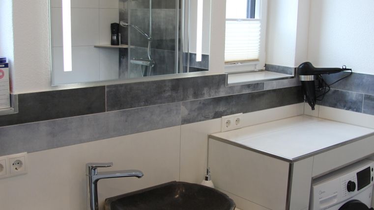 Une salle de bain moderne avec un lavabo en pierre naturelle et une machine à laver. Il y a également un grand miroir et une douche dans le coin.