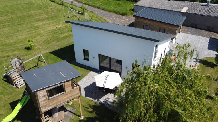 Une maison moderne, blanche avec un toit plat dans un environnement verdoyant. Au premier plan, il y a une tour de jeux avec un toboggan.