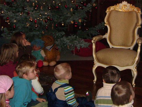 Eine Gruppe von Kindern sitzt vor einem festlich dekorierten Weihnachtsbaum und schaut auf einen leeren, eleganten Stuhl. Im Hintergrund steht ein Teddybär und es herrscht eine gemütliche Atmosphäre.
